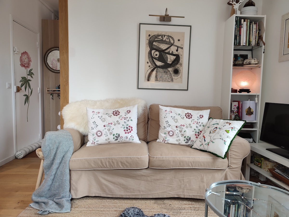 A cozy living area features a comfortable beige sofa adorned with floral cushions. A soft throw drapes over the side. A glass coffee table sits in front of the sofa, while a piece of abstract wall art adds interest. Shelving with decorative items is visible in the background.