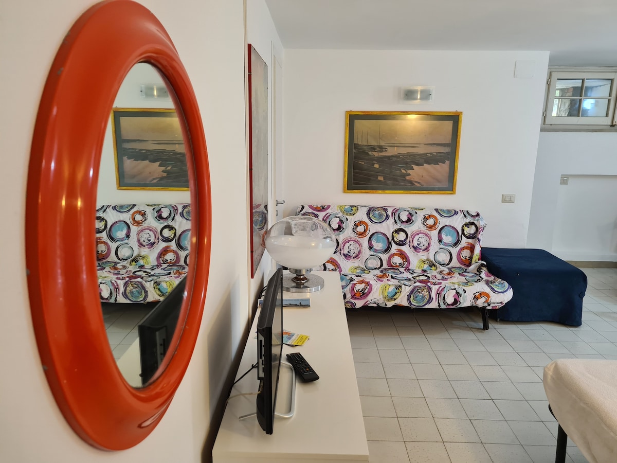 A bright living area is shown with a colorful patterned sofa that offers ample seating. A large round mirror reflects light, while a sleek TV stands on a white console. Art pieces with coastal themes complement the tranquil ambiance of the space.