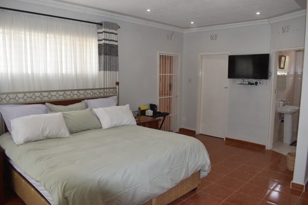 A spacious sleeping area features a large bed with multiple pillows and light green bedding. Natural light floods the room through large curtains, while a small desk with a coffee maker is positioned nearby. A wall-mounted television and an en-suite bathroom are also visible.