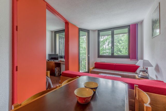 A cozy living area features bright red accents, including a sofa area and folding doors that provide access to a bedroom. A wooden dining table is set with bowls, while large windows invite natural light and offer views of greenery outside.