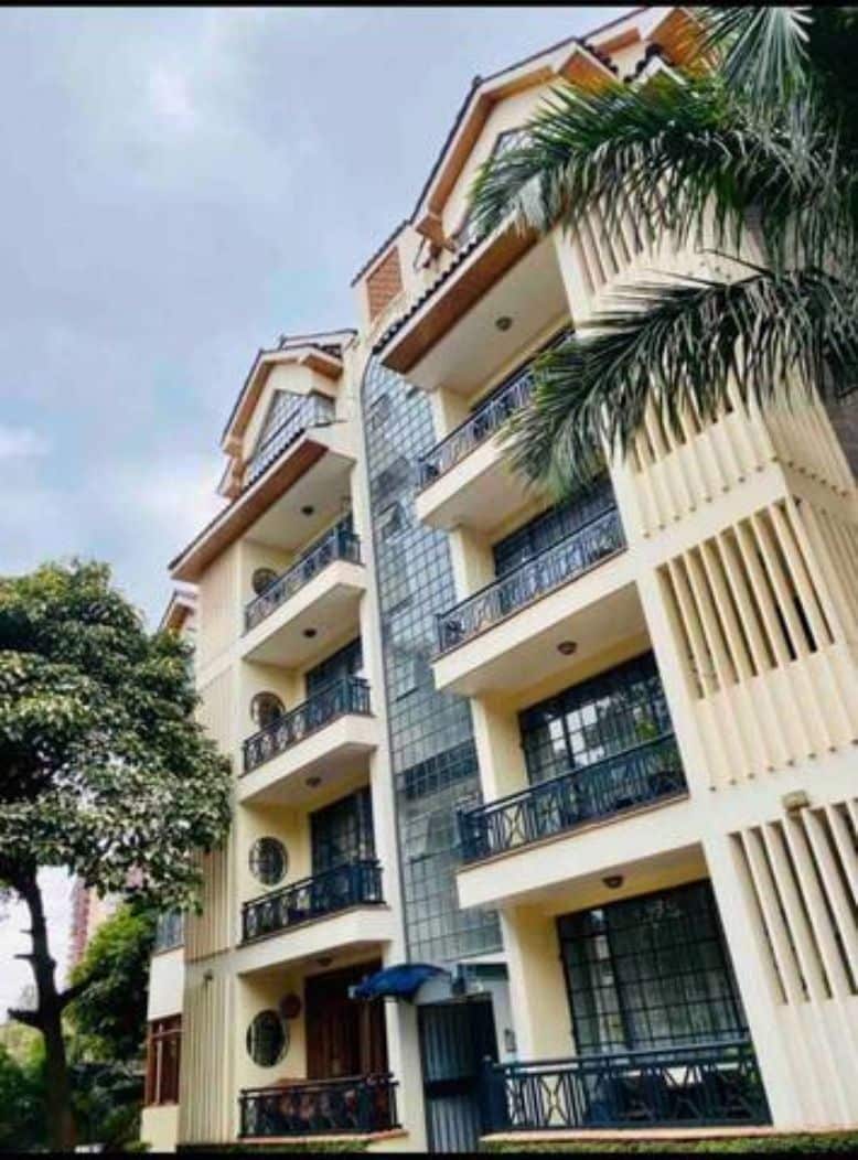 The exterior of the apartment building is shown, featuring multiple balconies with iron railings. Tall trees and lush greenery frame the structure, which consists of three floors. Large windows allow natural light to enter, enhancing the building's architectural features.