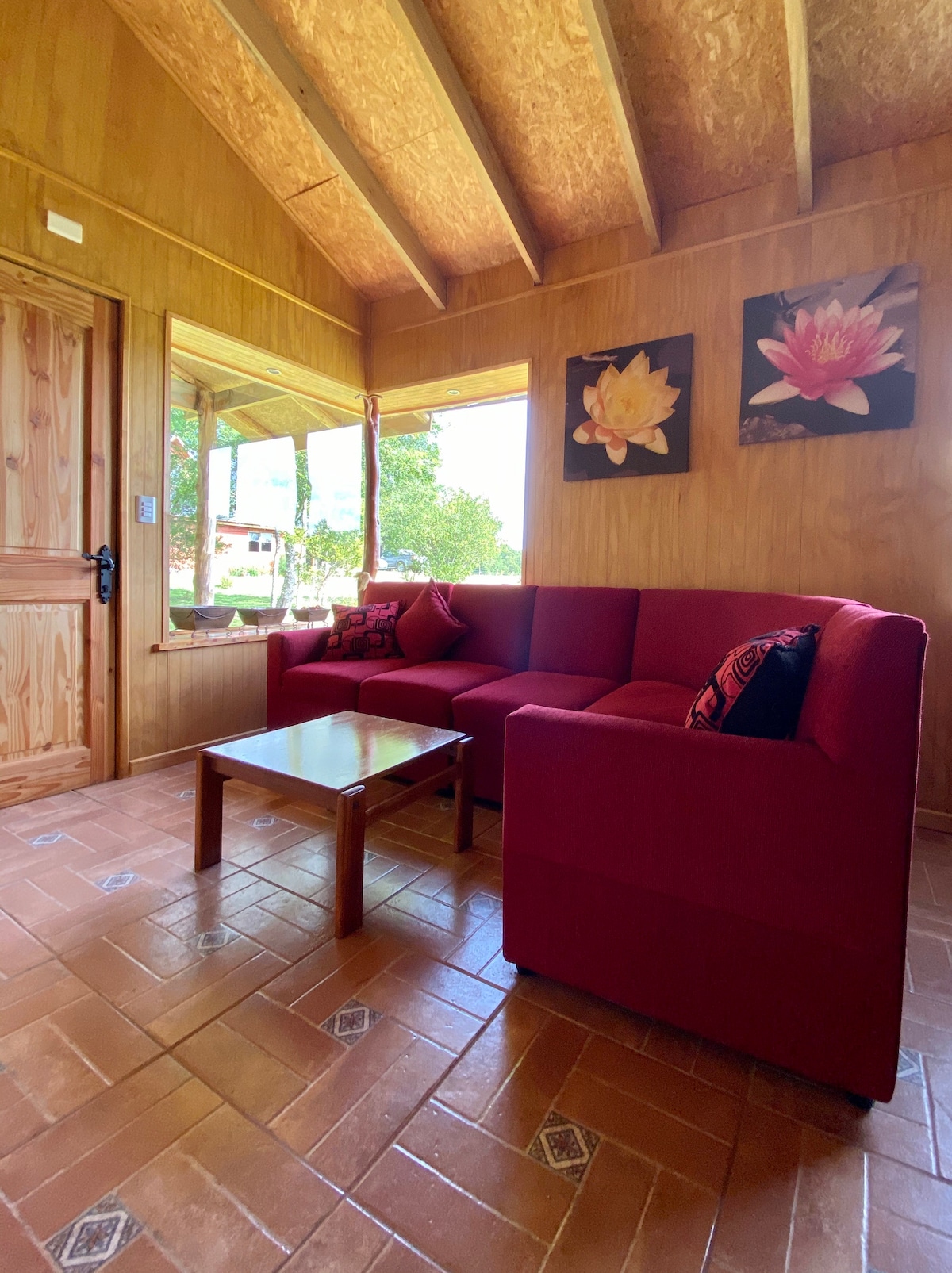 A living area features a red sofa, accented with decorative pillows. A small wooden table is situated in front, while large windows allow natural light to fill the space and offer views of the surrounding greenery. Floral artwork adorns the walls.