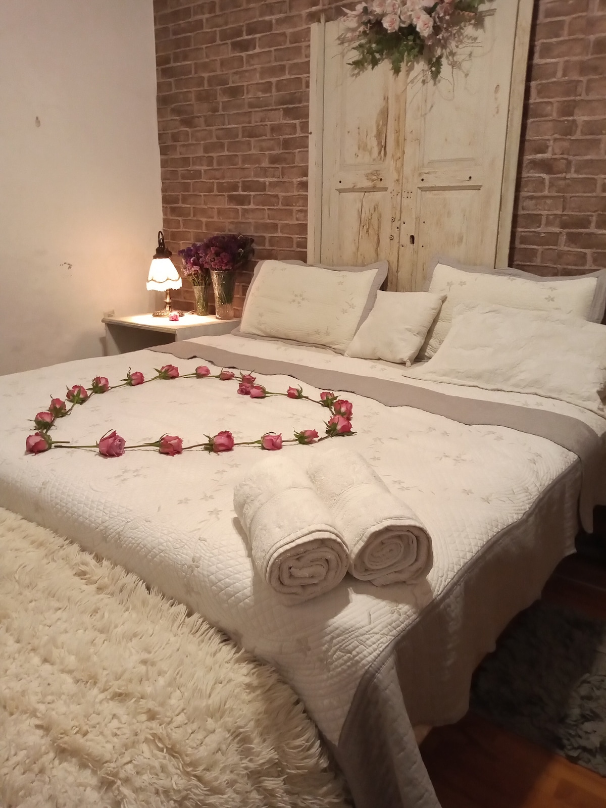 A private bedroom is depicted with a large bed, adorned with a decorative arrangement of roses. Fresh towels are neatly placed at the foot of the bed. Soft textures from the bedding and plush area rug enhance comfort, while warm light emanates from a bedside lamp.