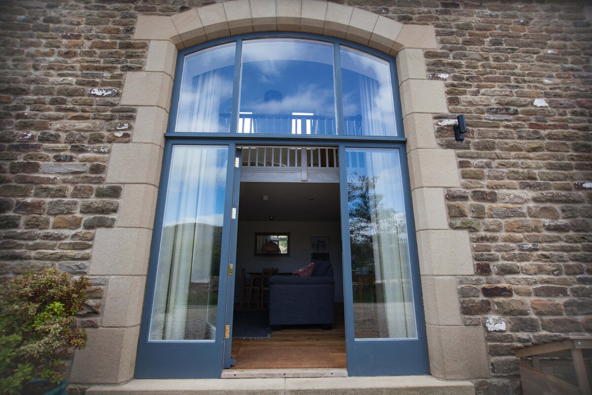 A striking entrance showcases large, full-height barn door windows framed in a deep blue finish. The stone exterior of the cottage is visible, providing a rustic charm. The interior can be glimpsed, revealing a comfortable living area beyond the threshold.