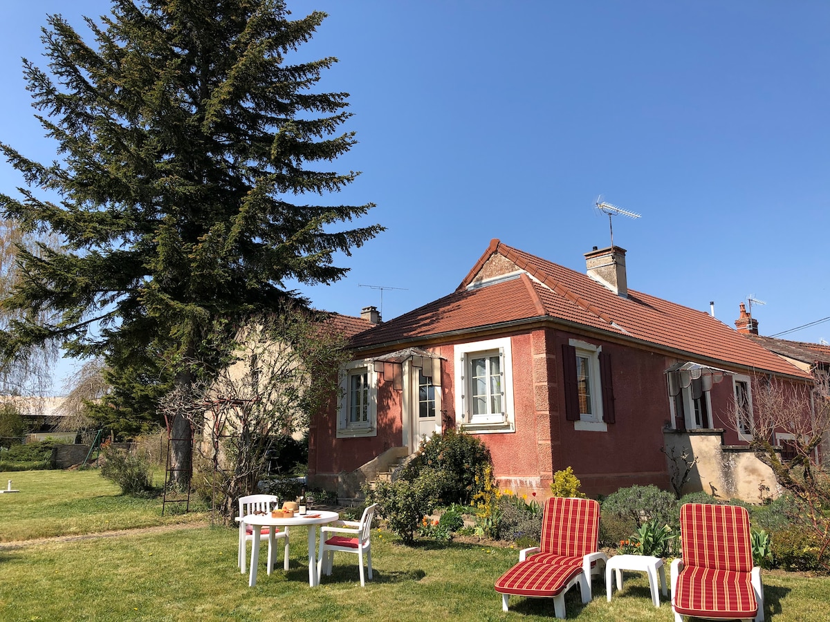 A charming red house is surrounded by a well-maintained garden, featuring a large tree and green grass. Outdoor seating is provided with two lounge chairs and a table, offering a serene space for relaxation under the clear blue sky.