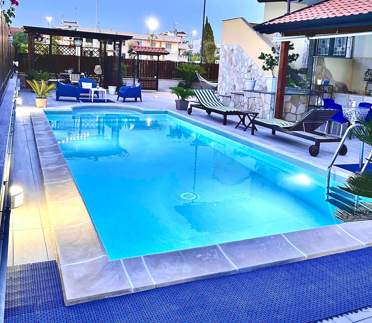 A serene outdoor space features a crystal-clear swimming pool surrounded by comfortable loungers and tropical plants. The area is illuminated softly in the evening light, creating a relaxing ambiance. A stylish gazebo can be seen in the background, providing additional seating options.