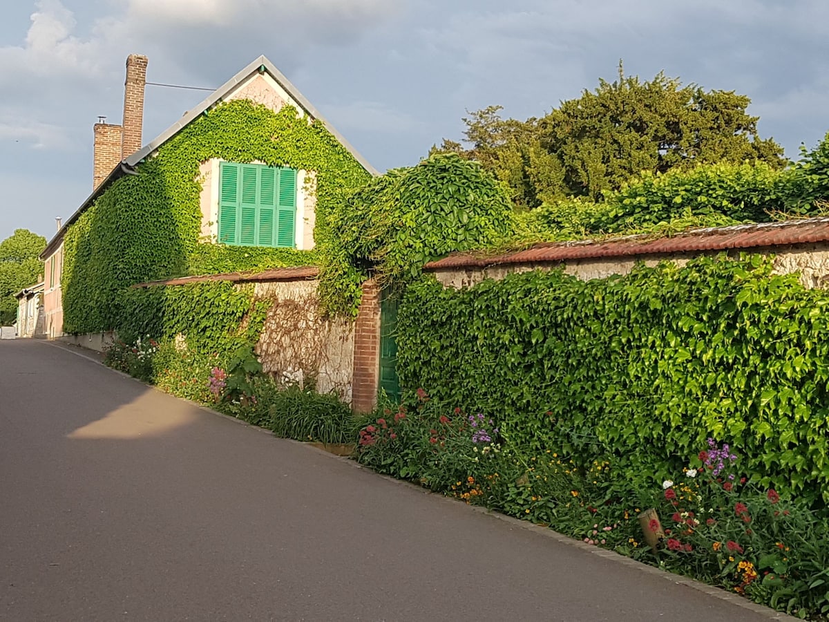 A quaint house is enveloped in lush green ivy, with a striking blue window adding a pop of color. The surrounding garden features vibrant flowers, contributing to a serene and inviting atmosphere. A quiet street can be seen alongside the property.