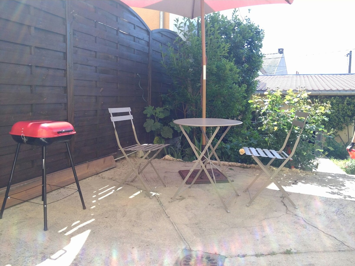 A small outdoor terrace features a round table with two folding chairs under a red parasol. A charcoal barbecue stands nearby, surrounded by greenery and wooden fencing, creating a private space for outdoor dining and relaxation.