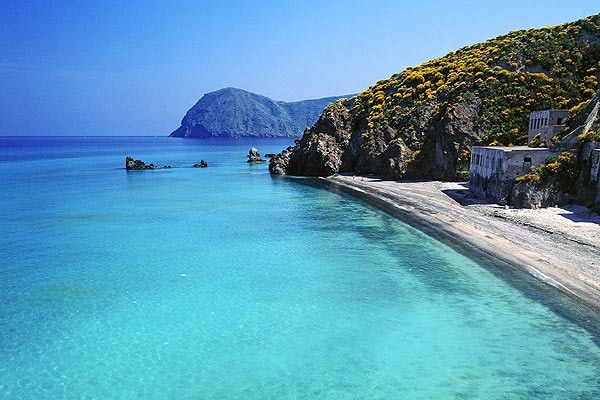 A serene coastline is presented, featuring soft sandy beach bordered by clear turquoise waters. Lush green hills rise in the background, creating a natural backdrop. Rocky formations are visible along the shore, adding texture to the landscape.