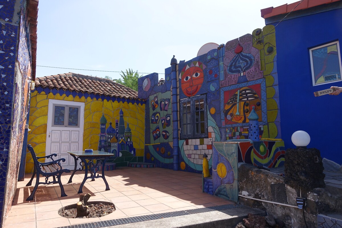 An outdoor area showcases vibrant mosaics and murals on the walls, featuring various playful designs. A small seating arrangement with two chairs and a table is visible, complemented by an assortment of colorful artistic elements and a sunlit tiled floor.
