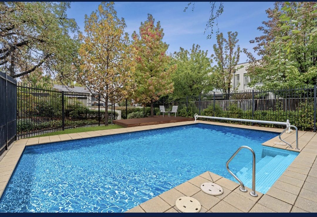A swimming pool is framed by greenery, featuring clear blue water and surrounded by a tile deck. Sunlight reflects off the surface, with trees and shrubs providing a serene backdrop. A safety railing and steps are visible, enhancing accessibility.