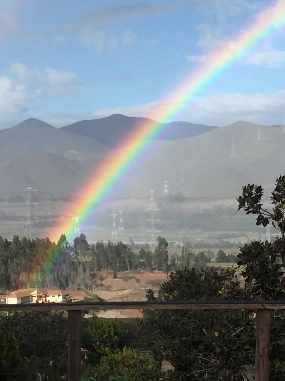 A vibrant rainbow arcs across a mountainous landscape, connecting with the ground in a lush area featuring greenery. Distant power lines run through the valley, while clouds hover gently in the sky, creating a serene backdrop for the colorful display.
