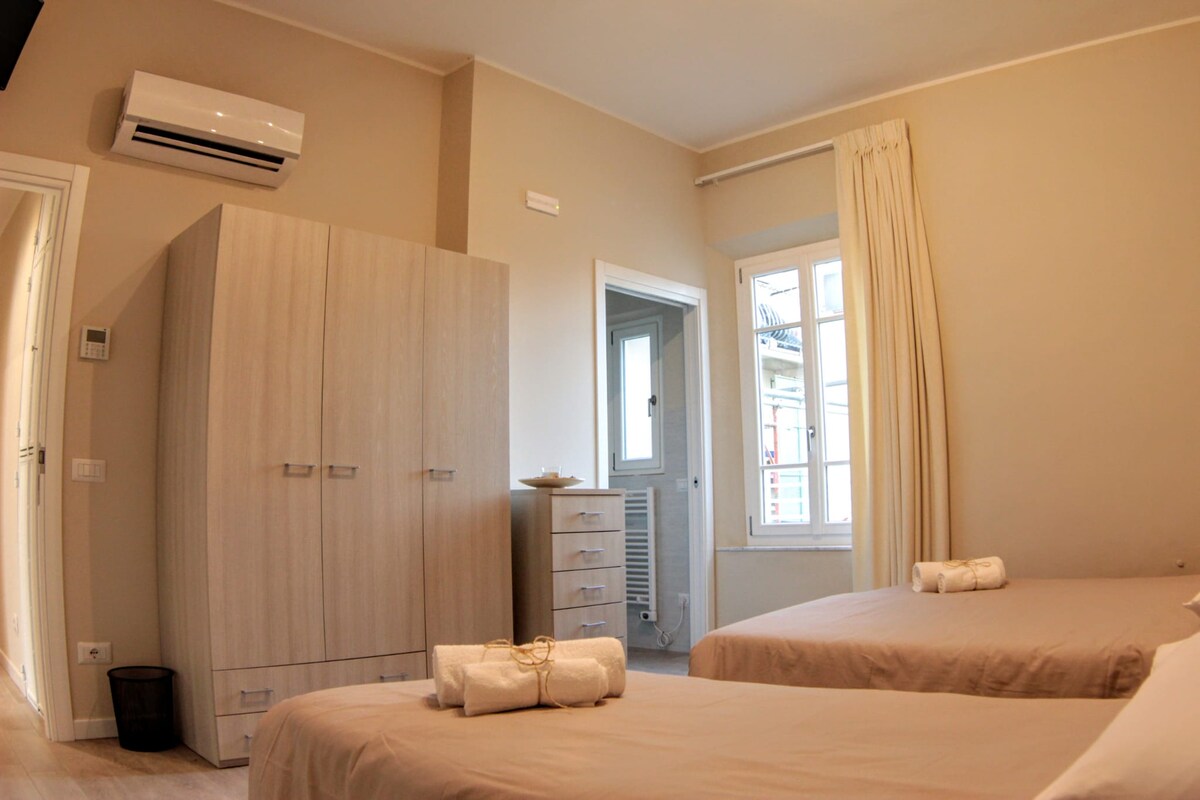 The room features two spacious beds covered with neutral linens. A wardrobe and a tall dresser provide ample storage. Natural light enters through a window, complementing the warm tones of the walls and furnishings. Wall-mounted air conditioning is visible for climate control.