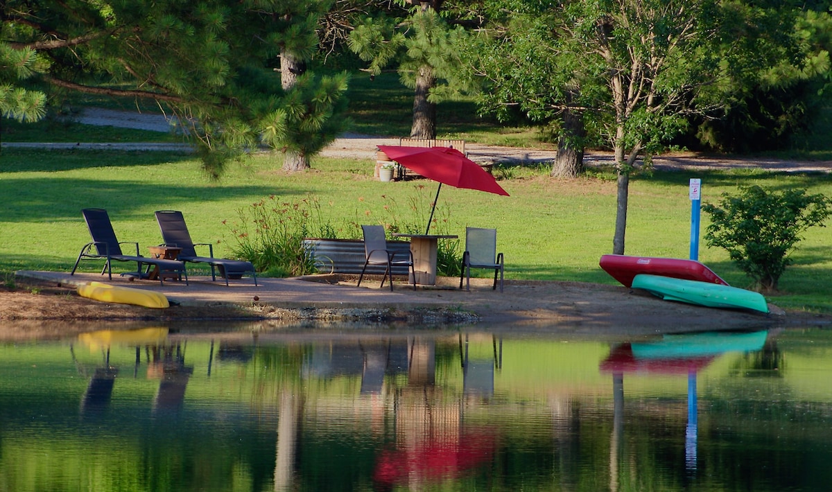 A serene beach area is visible by the pond, featuring a couple of lounge chairs and a red umbrella providing shade. Kayaks are positioned on the sandy shore, reflecting the lush greenery and calm water surface.