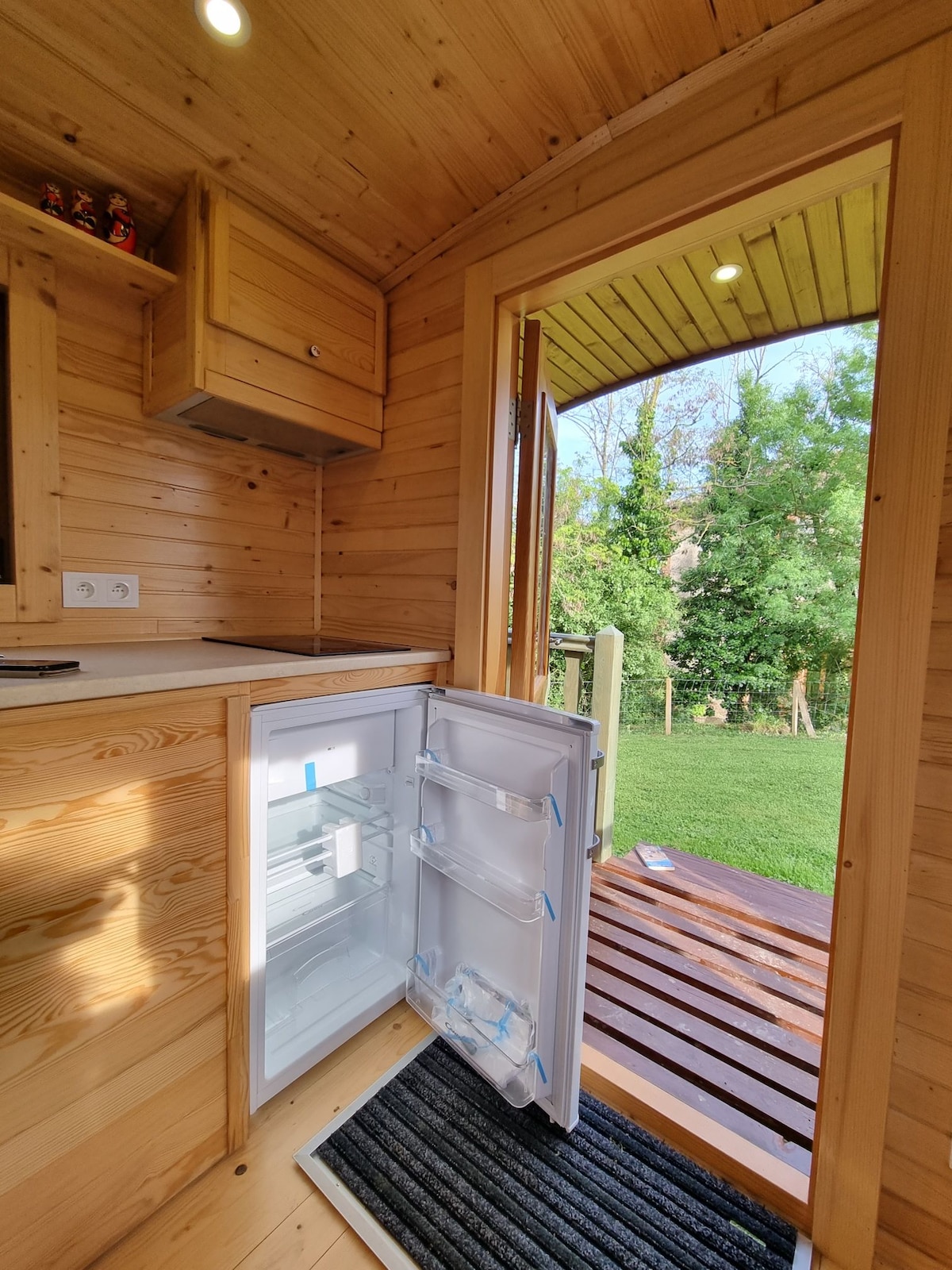 An open doorway reveals a view of the lush garden outside, framed by wooden walls. A refrigerator is visible, showcasing transparent shelves. Natural light enters, enhancing the warm tones of the wooden interior and inviting the outdoors inside.