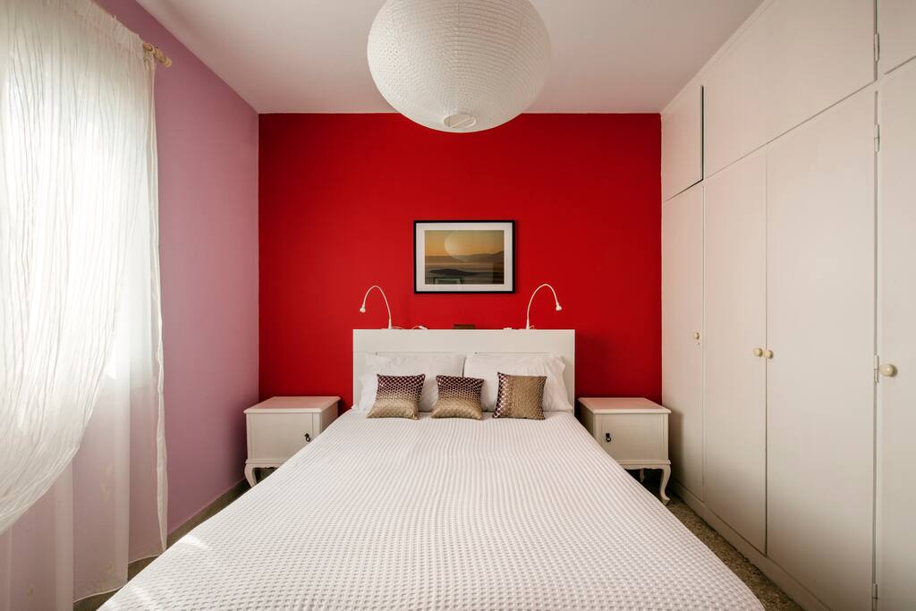 The bedroom in morning light. There is a big mirror, a TV, and a closet for hanging clothes or putting them in the drawers. The furniture by the bed and the bed back also offer storage spaces.