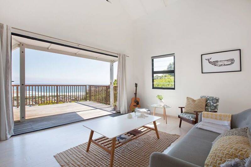A bright living area is showcased with large glass doors that open to a deck with ocean views. A comfortable gray sofa and a wooden coffee table are positioned on a light-colored rug. Natural light fills the space, enhancing the uncluttered, inviting environment.