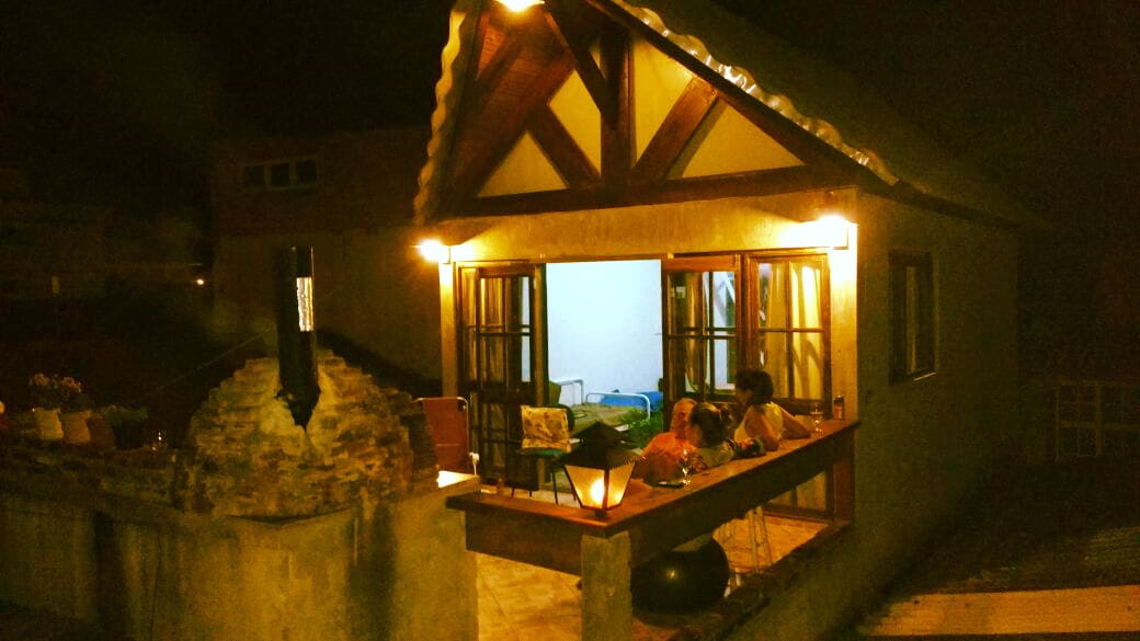 A charming exterior view of a two-story house is presented at night, featuring warm lights that illuminate the balcony area and patio. The stone barbecue is visible, complementing the inviting atmosphere created by open double doors leading into the living space with cozy seating.