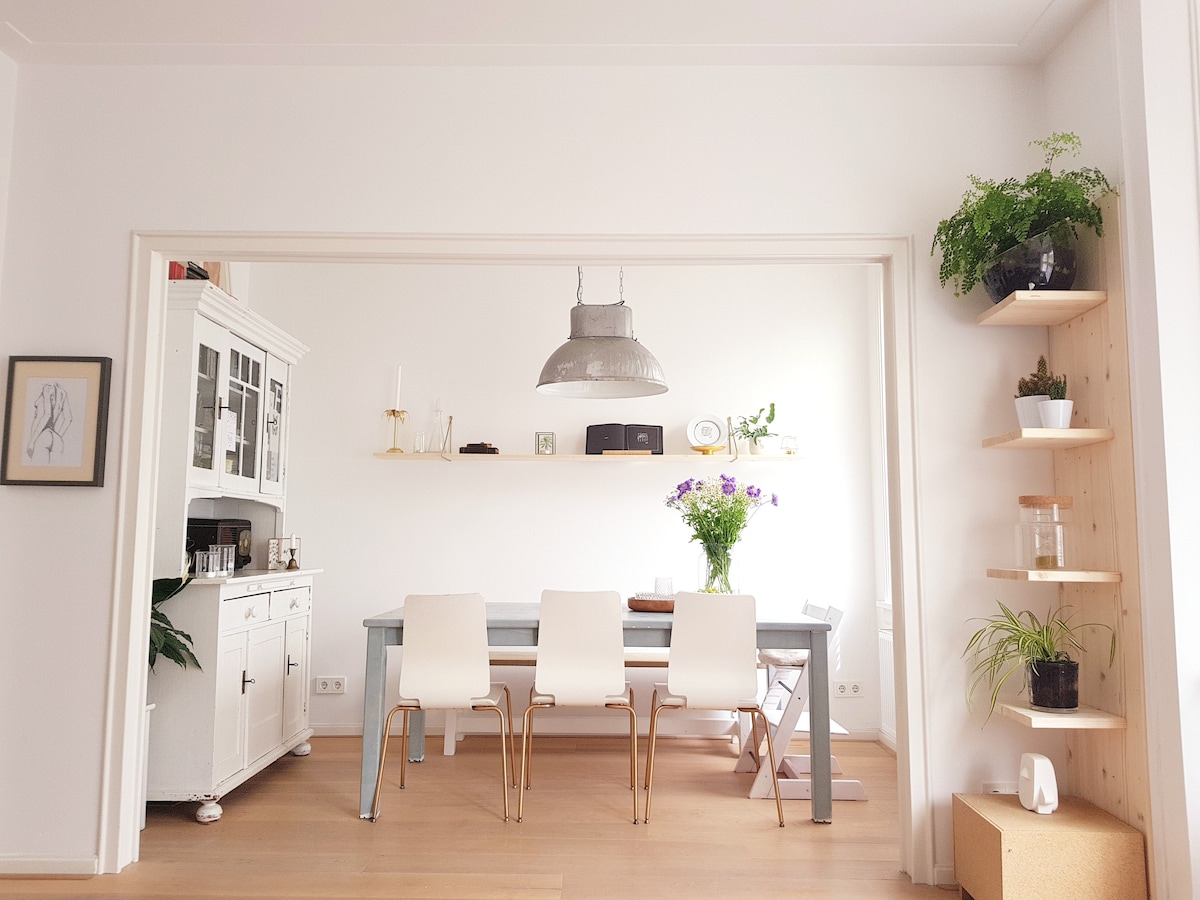 A well-lit dining area features a long table surrounded by four sleek white chairs. A modern pendant light hangs above the table. The walls are adorned with minimal decor, while a shelf holds potted plants in the corner, enhancing the bright ambiance.