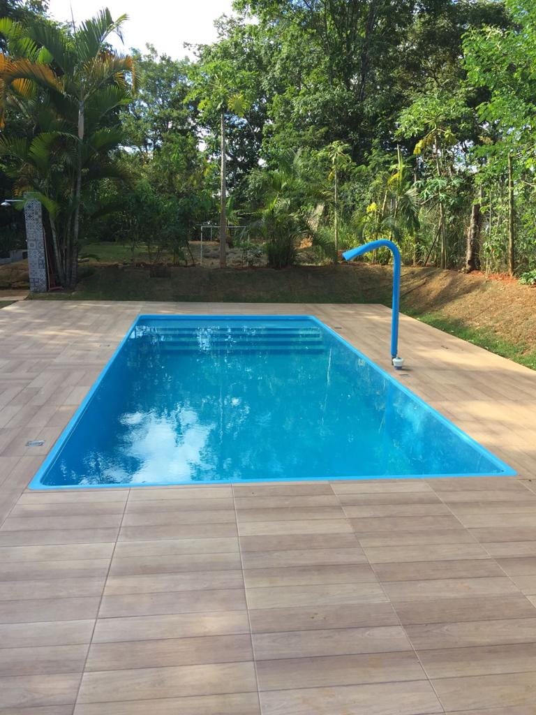 A clear blue swimming pool is set amid lush greenery, with a spacious wooden deck surrounding it. A water slide with a vibrant blue finish is positioned at the far end of the pool, inviting enjoyment and relaxation in a natural setting.