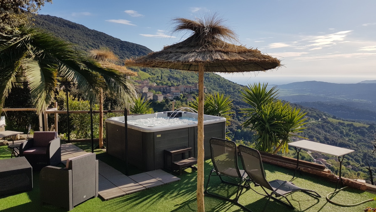 A private outdoor spa area is set against a panoramic landscape of rolling green hills and distant sea views. Comfortable seating is arranged under a thatched umbrella, with loungers positioned nearby. Lush palm trees enhance the serene atmosphere of the space.