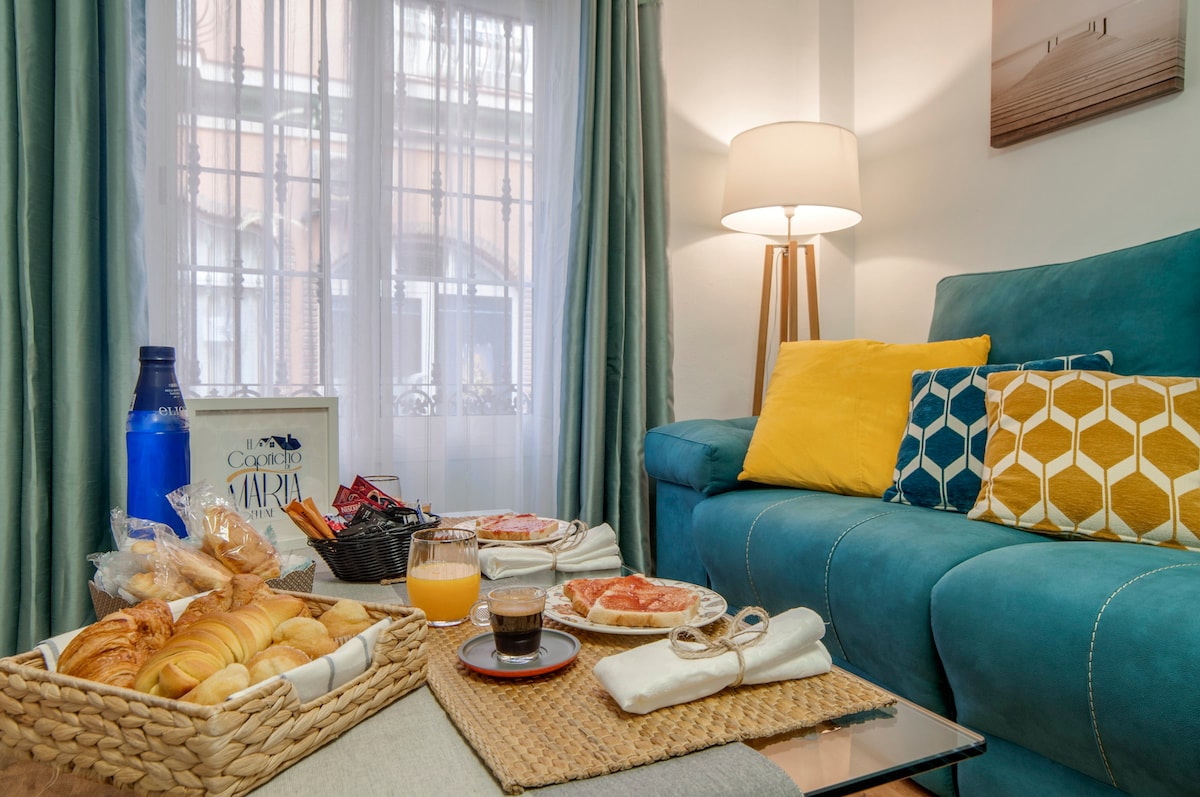 A welcoming living area features a comfortable teal sofa adorned with vibrant yellow and patterned cushions. A small table displays a selection of pastries, orange juice, and coffee. Natural light filters through sheer curtains, creating a bright and inviting space.