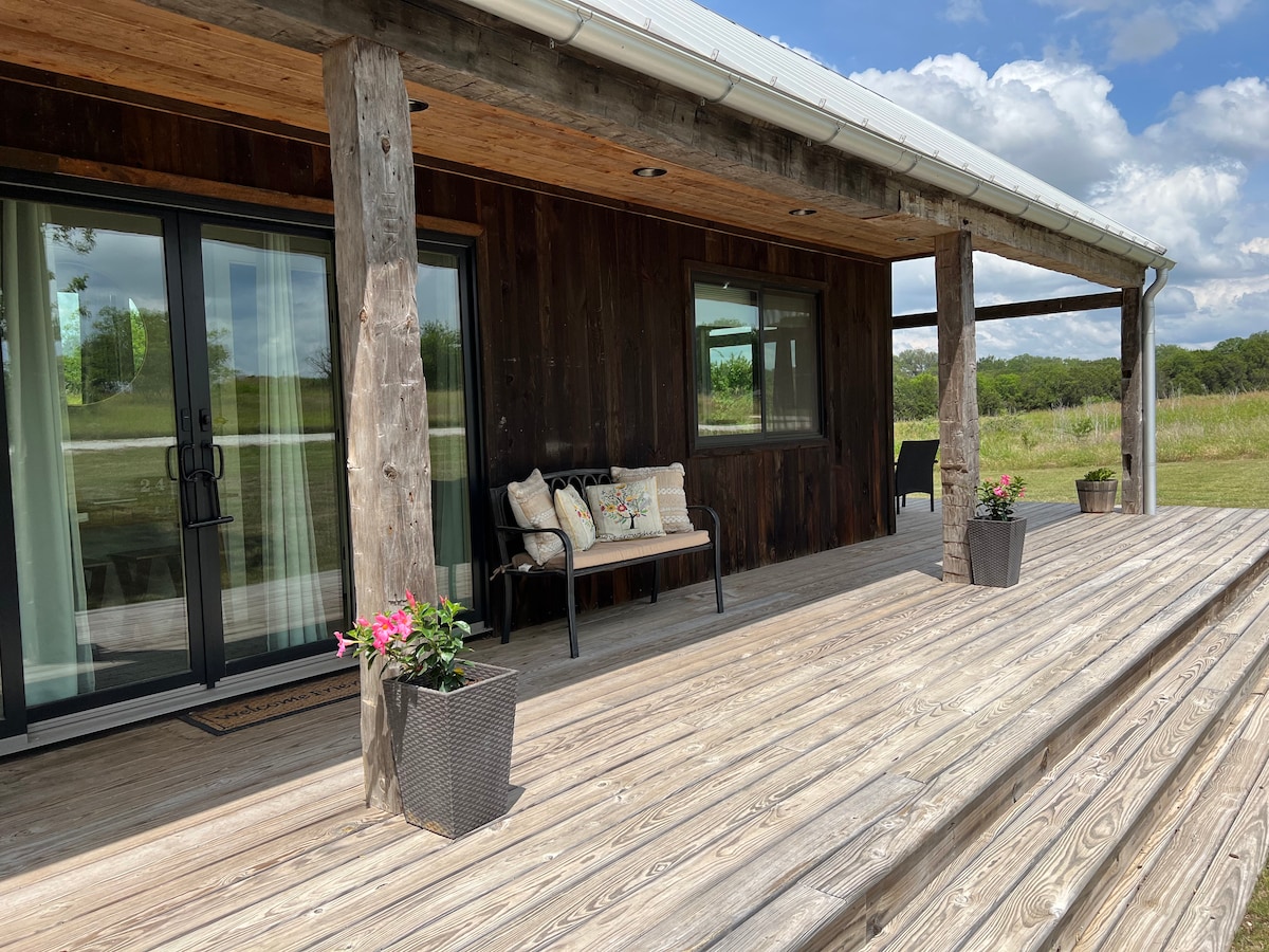 A spacious wrap-around deck features wooden flooring and is accented by potted flowers. A seating area with a bench and cushions is positioned beside large glass sliding doors, offering a view of the surrounding open fields and blue sky.