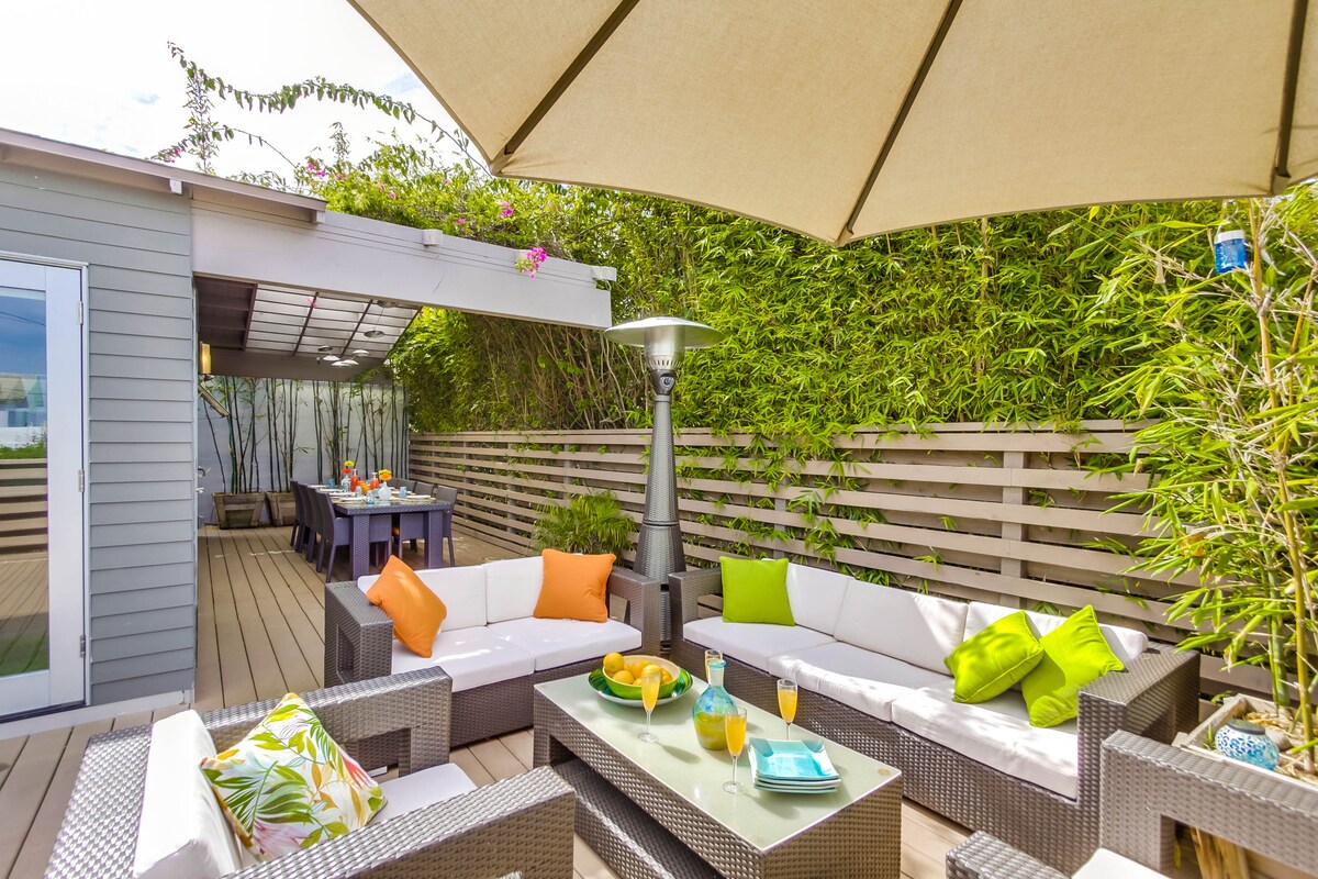 An outdoor deck area is presented with comfortable seating featuring colorful cushions. A large, shaded dining table is visible in the background, surrounded by lush bamboo greenery. A small table in the foreground holds drinks and fresh fruit, enhancing the inviting, relaxing environment.