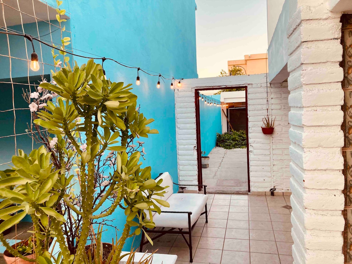 A serene outdoor area is framed by vibrant blue walls. String lights add a soft glow overhead, and potted plants bring greenery to the space. A comfortable seating option is positioned near a doorway leading to a pathway, enhancing the inviting atmosphere.