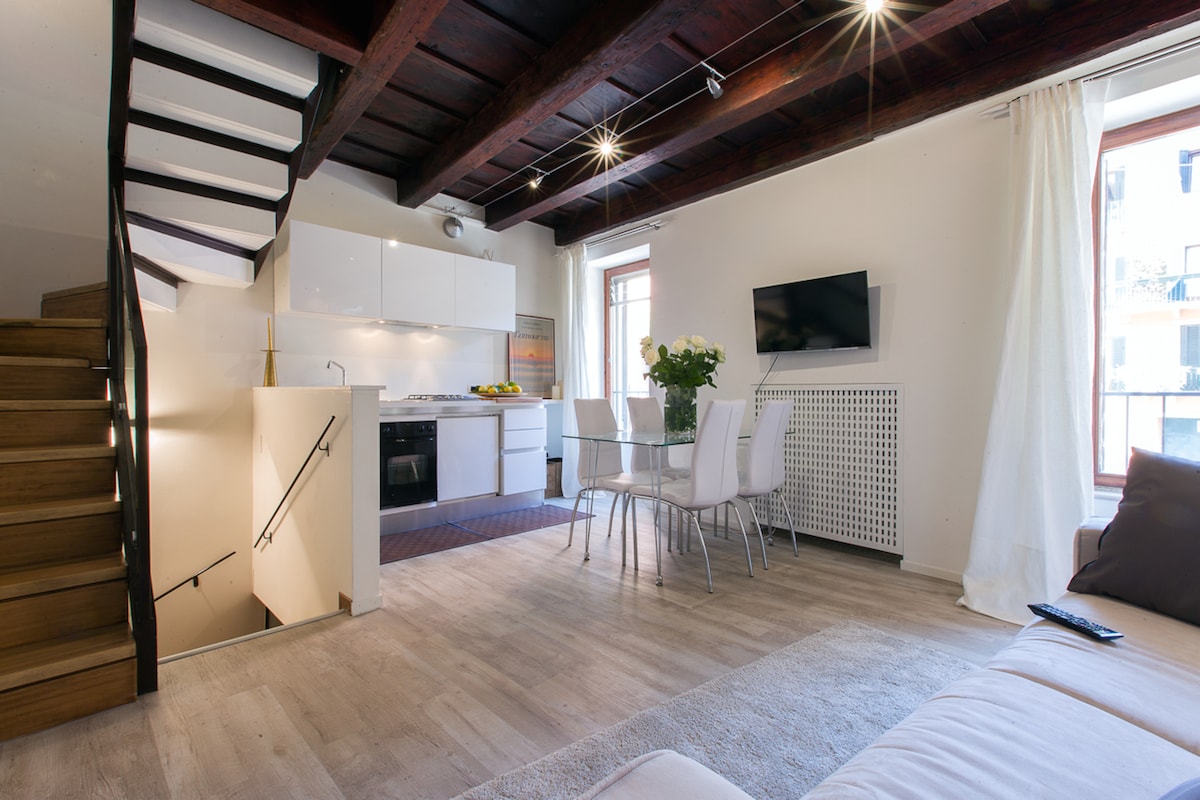 An open-plan living area features a modern kitchen with white cabinetry and a dining table with six chairs. Natural light fills the space through large windows. A stylish staircase leads to the upper level, while a cozy lounging area is equipped with a comfortable sofa.