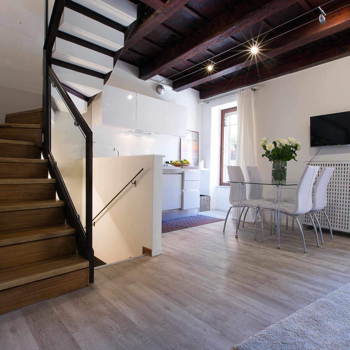 A modern living space featuring a wooden staircase leading to the upper level. The kitchen appears equipped with sleek cabinetry and counter space. A dining area showcases a table with white chairs, while a bouquet of flowers adds a touch of vibrancy. Natural light fills the room through the window.