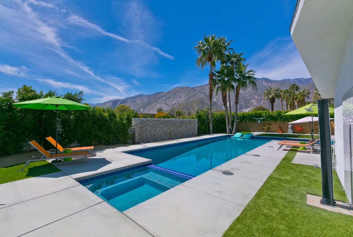 A clear view of a resort-style saltwater pool is presented, bordered by vibrant green palm trees. Loungers in bright colors provide seating options, while a parasol offers shade. The backdrop features the San Jacinto Mountain, enhancing the outdoor experience.
