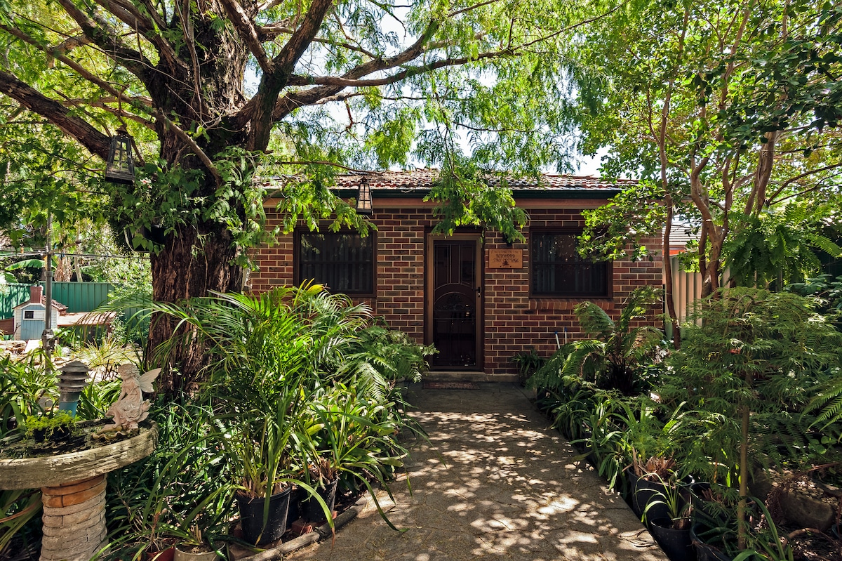The entrance to the guest cottage is framed by lush greenery, including ferns and potted plants. The red brick exterior of the cottage is visible, along with a secure door set beneath a canopy of leaves, creating a welcoming and peaceful entryway.