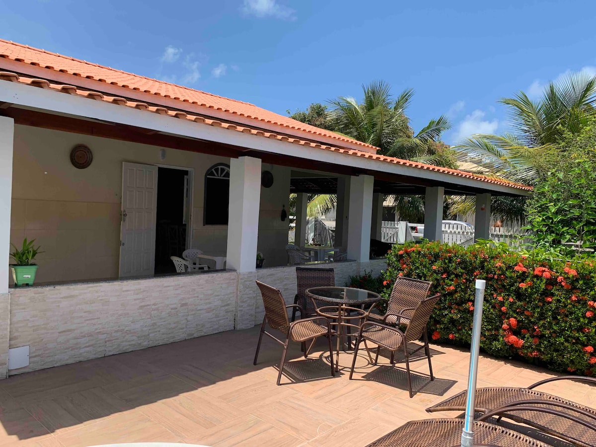 A well-maintained beach house features a large covered veranda with several outdoor seating options. The porch is surrounded by lush greenery and colorful flowers, creating a serene environment. The architecture includes a tiled roof and multiple windows for natural light.
