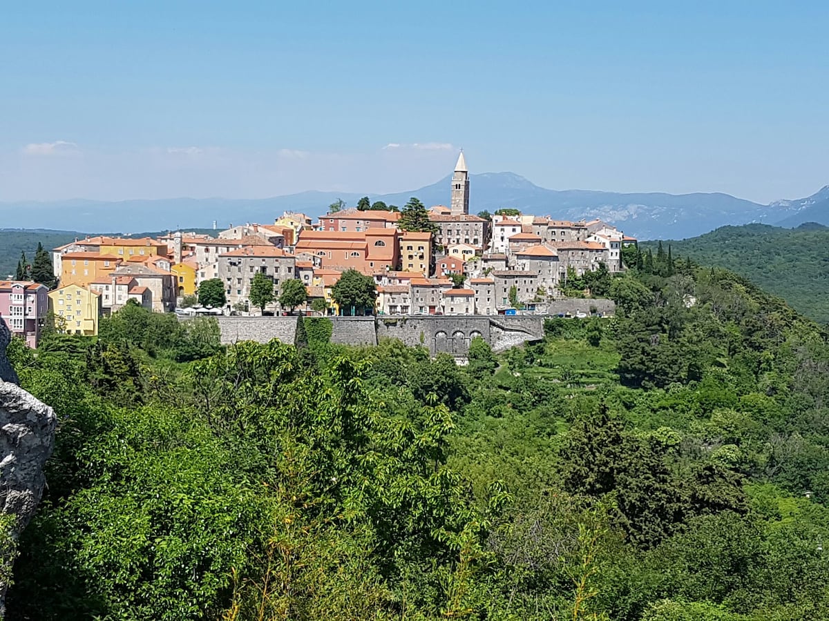 A panoramic view of Labin old city is captured, showcasing its terracotta-roofed buildings and a prominent bell tower. Green hills surround the city, providing a natural backdrop. The clear blue sky enhances the scenery, highlighting the charm of this historic area.