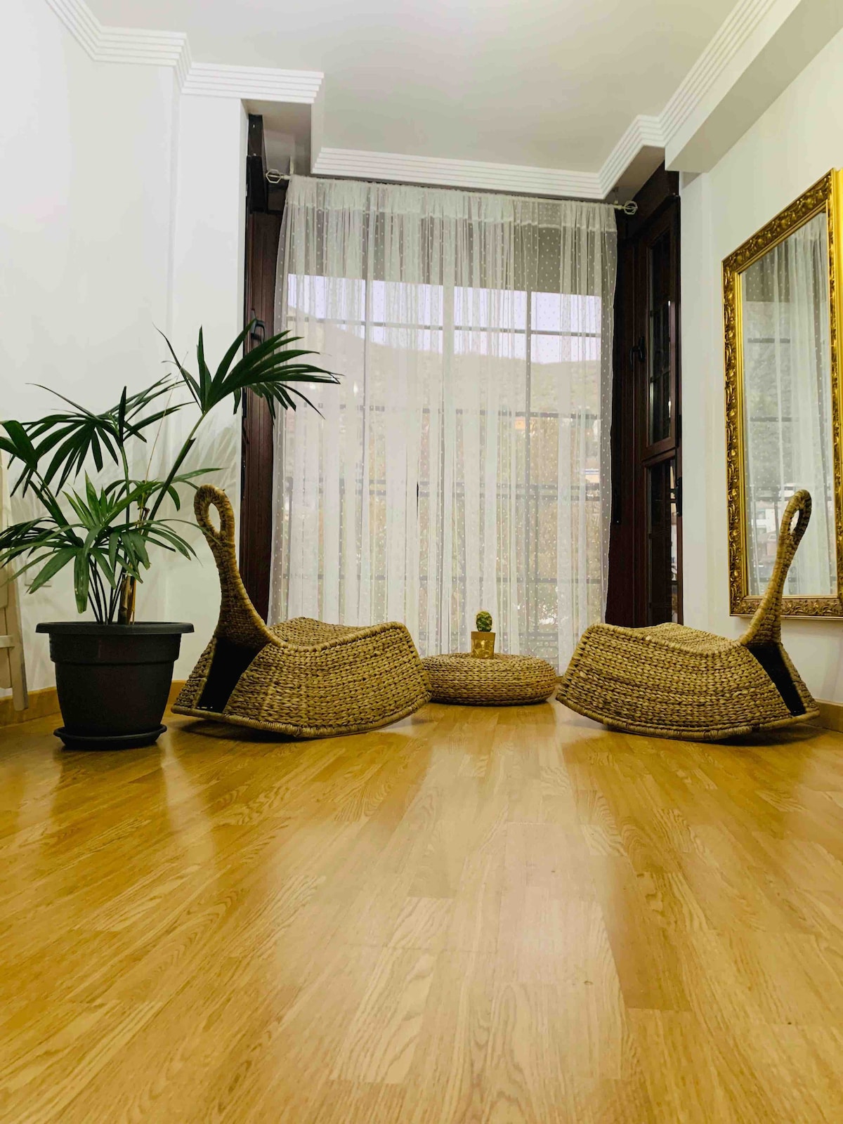 A bright seating area features two woven chairs facing a small circular table, arranged on light wood flooring. Natural light filters through sheer curtains, creating a serene environment. A potted plant adds a touch of greenery beside a tall mirror, enhancing the room's spacious feel.