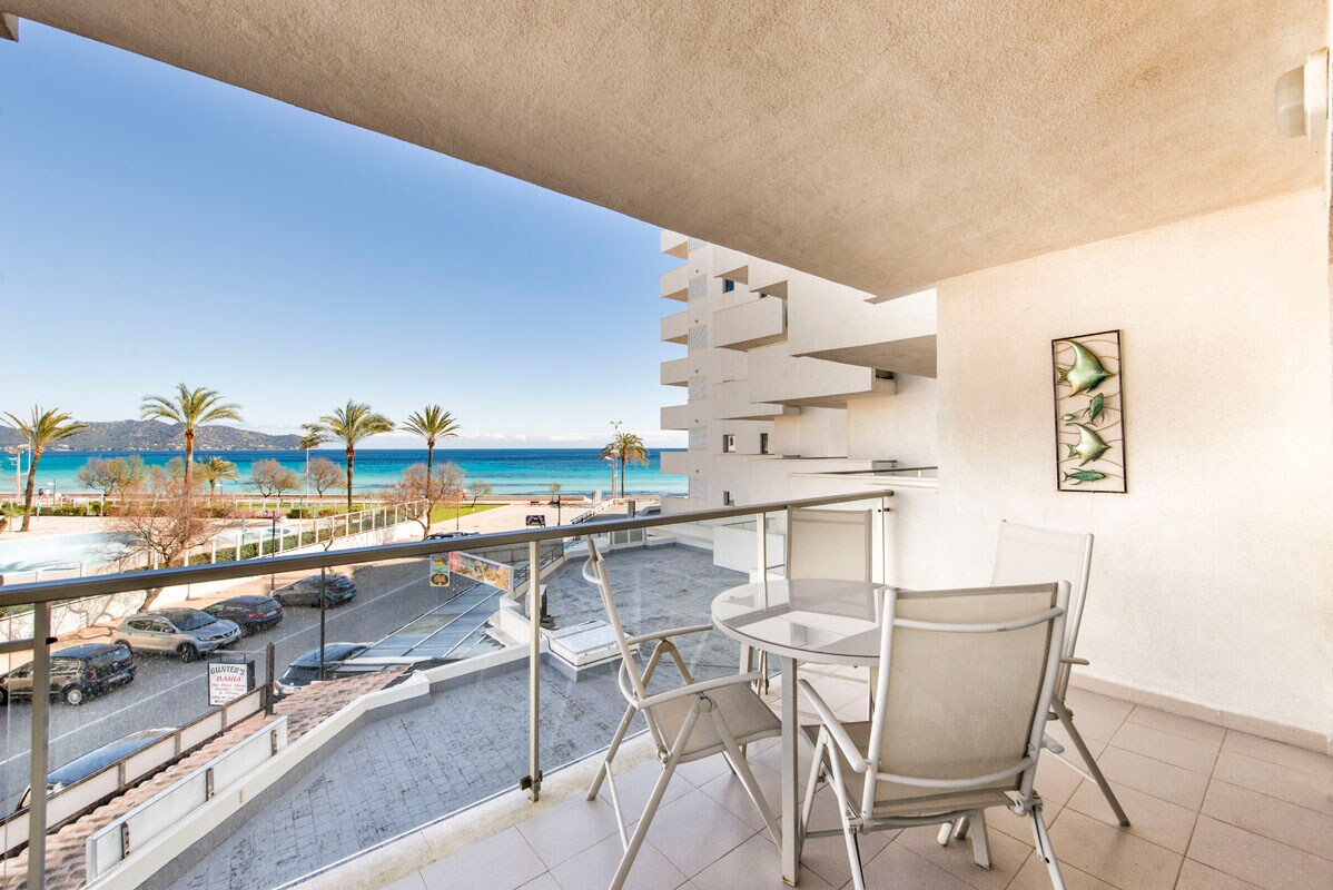 A balcony offers a clear view of the sea, complemented by palm trees lining the beach. Minimalist outdoor furniture, consisting of two chairs and a small table, is arranged on the tiled surface. The surrounding building features a light-colored exterior.