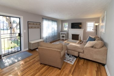 A bright living room features new hardwood flooring and large windows, allowing natural light to fill the space. A cozy sectional sofa is positioned for comfort, while a small coffee table sits in front of a television mounted above the fireplace, creating an inviting atmosphere.