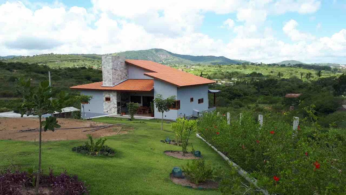 A modern building is positioned on a lush, green landscape, surrounded by gentle hills. The structure features a distinct sloped roof and a combination of stone and stucco materials. Trees and neatly arranged plants enhance the exterior, creating a serene environment.