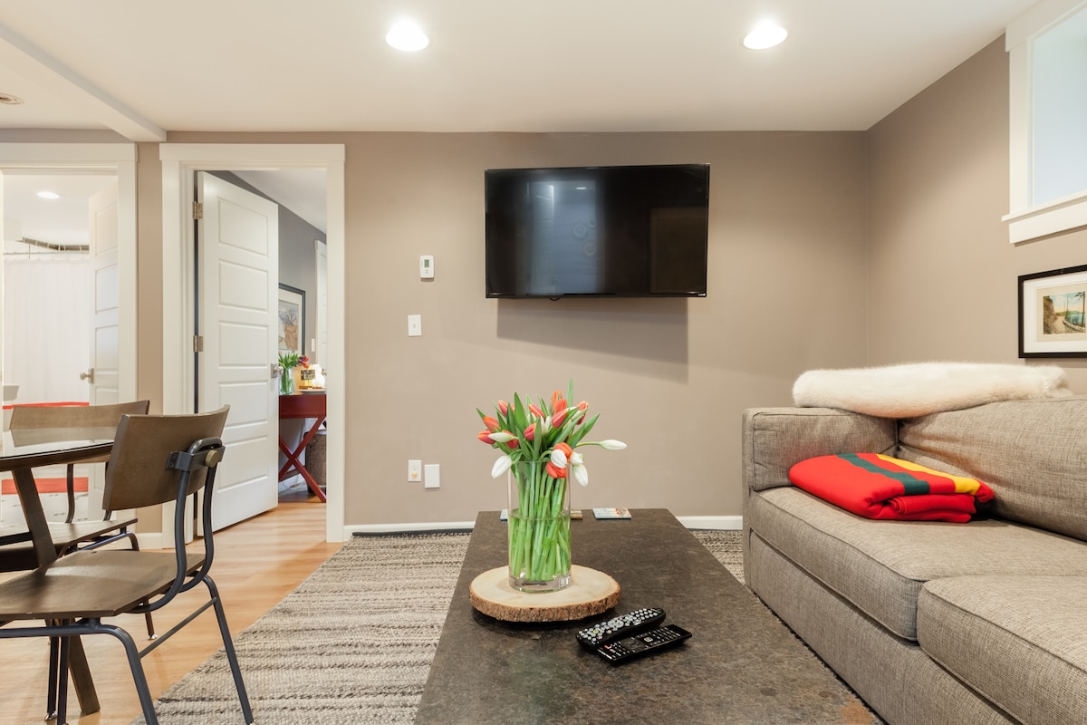 A comfortable seating area is presented with a gray sofa and a circular coffee table adorned with a vase of tulips. A flat-screen TV is mounted on the wall. The dining space is visible through a doorway, featuring a red table set for four.
