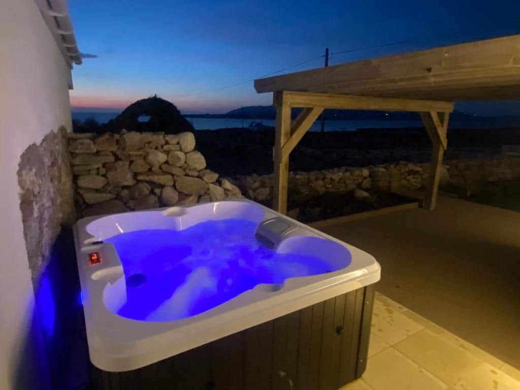 An outdoor hot tub is illuminated with soft blue lighting, set against a backdrop of a twilight sky. A wooden pergola provides shelter, while stone walls add rustic charm. The serene coastal views are visible in the distance, enhancing the relaxing atmosphere.