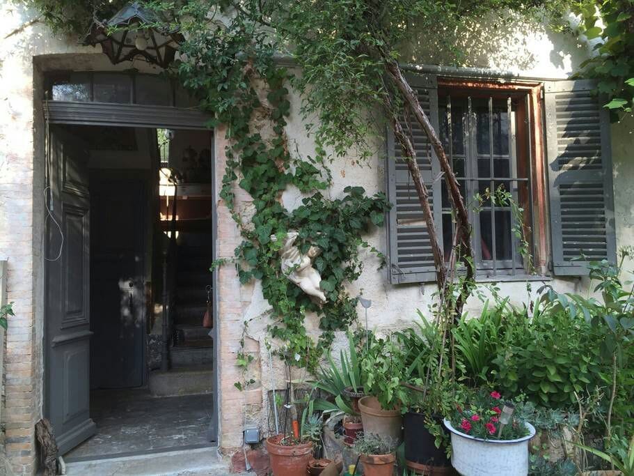 The exterior entrance is framed by lush greenery and flowering plants. A statue can be seen on the wall, adding character to the rustic facade. Light streams through the open window, highlighting the inviting doorway and the staircase leading inside.
