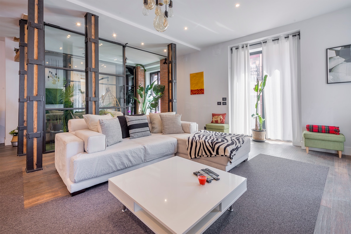 A spacious living area features a large white sectional sofa with accented cushions, complemented by a modern coffee table and a patterned rug. Natural light enters through sheer curtains, illuminating plants in the background, and a cozy reading nook is visible in the corner.