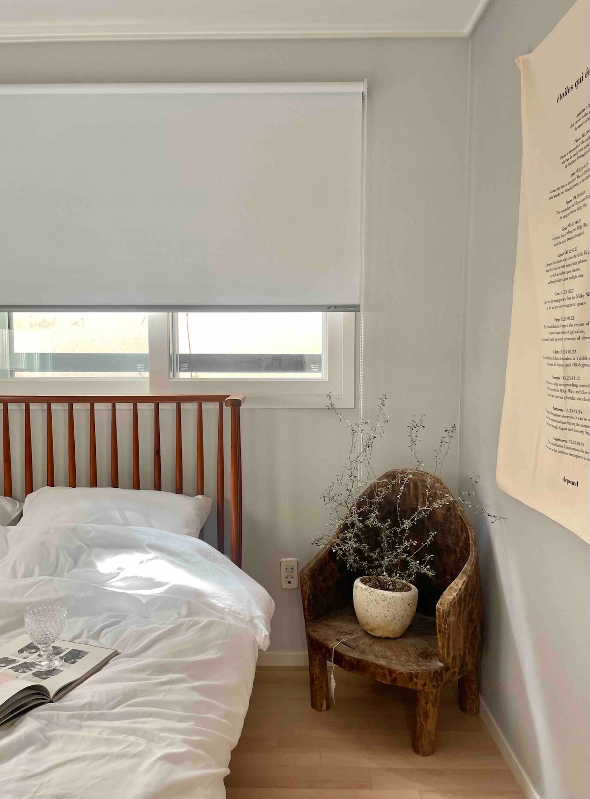 A serene bedroom features a wooden bed frame with white bedding, positioned beside a wall adorned with text art. A small, rustic chair holds a potted plant, adding an organic touch. Natural light streams in through a nearby window, covered by a light roller shade.