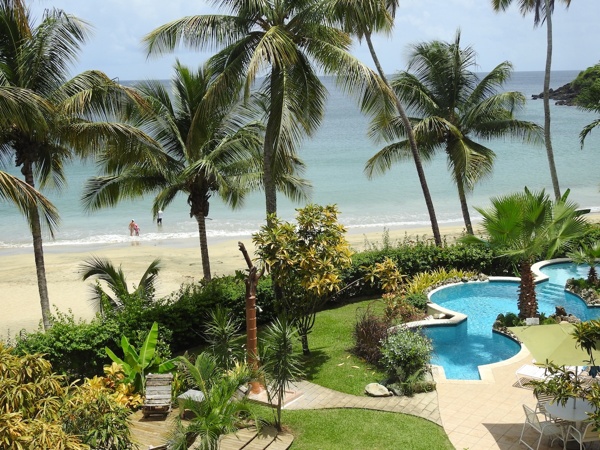 A view from the patio reveals a sandy beach lined with palm trees, leading to calm waters. The vibrant green landscaping frames a swimming pool, which is surrounded by lounge chairs. Sunlight casts soft shadows across the scene, enhancing the tranquil atmosphere.