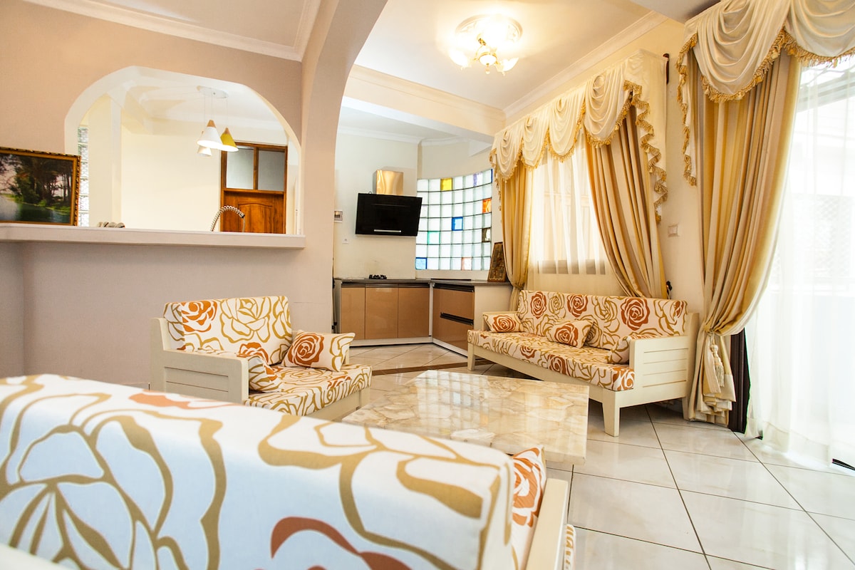 A living area features elegant furnishings with floral-patterned upholstery. Two upholstered chairs and a spacious sofa are arranged for comfortable seating. Natural light filters through large windows adorned with soft drapes. A TV is mounted on the wall, and a sleek kitchen area is visible in the background.