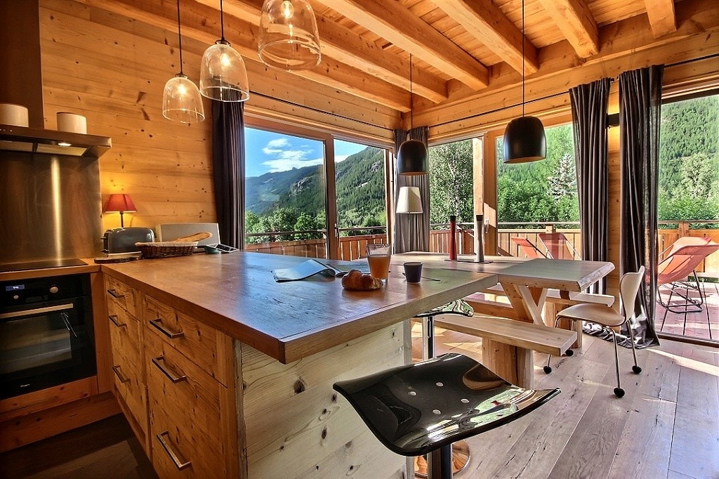 A modern kitchen with a wooden island is highlighted by large windows that frame a mountain view. Pendant lights hang above the island, and a dining table with seating is positioned nearby. Natural light enhances the warm wooden interiors, creating an open and inviting space.