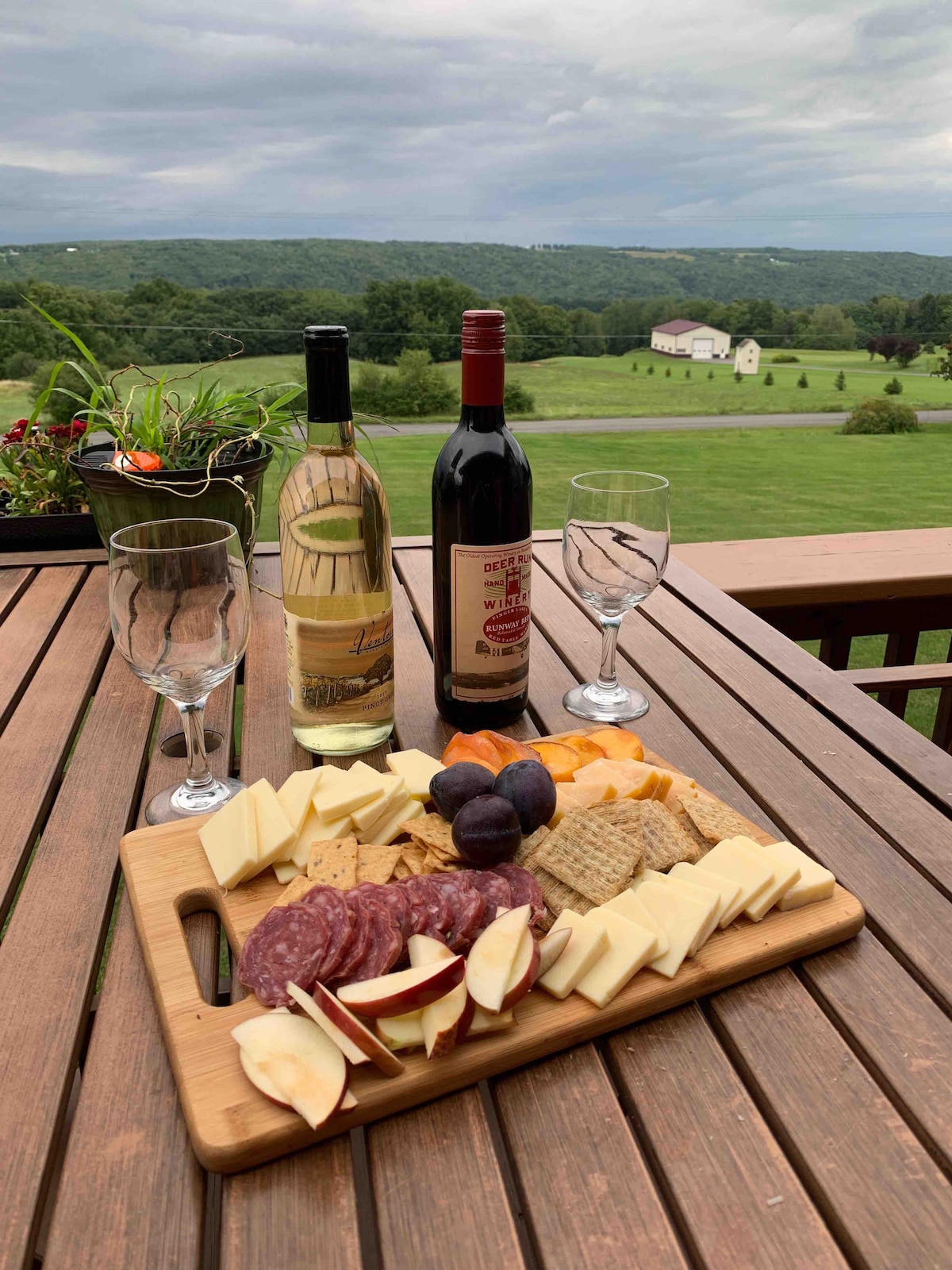 A wooden table is set outside, featuring a charcuterie board with cheese, meats, fruits, and crackers. Two bottles of wine and four glasses are arranged beside the board. A panoramic view of rolling hills and a distant house is visible in the background.