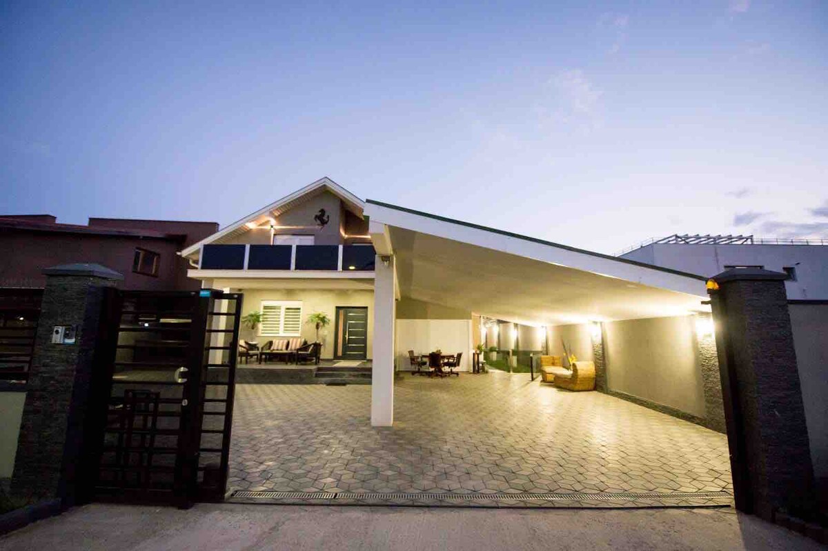 The entrance to the villa is showcased, featuring a spacious covered carport with lighting enhancing the evening ambiance. A pathway leads to the main entrance, framed by greenery. Outside seating is visible, inviting relaxation in the tranquil setting.