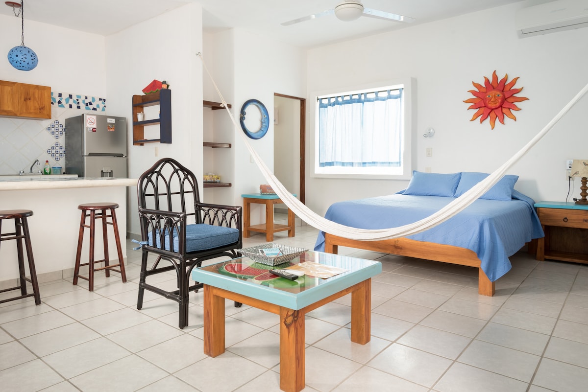 A bright and airy living area features a spacious bed draped in blue linens. A hammock is suspended between two posts, inviting relaxation. A dining table and stools are positioned nearby, with colorfully accented decor enhancing the atmosphere. Natural light fills the room through a window adorned with light curtains.